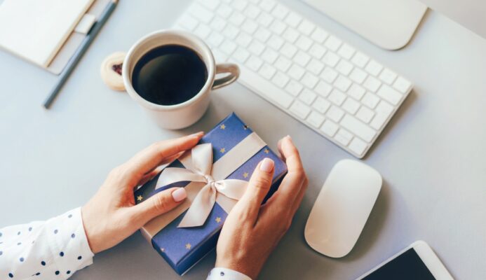 Nhân viên văn phòng mở quà tặng doanh nghiệp bên cạnh máy tính và tách cà phê, mang lại không khí thân thiện nơi làm việc.